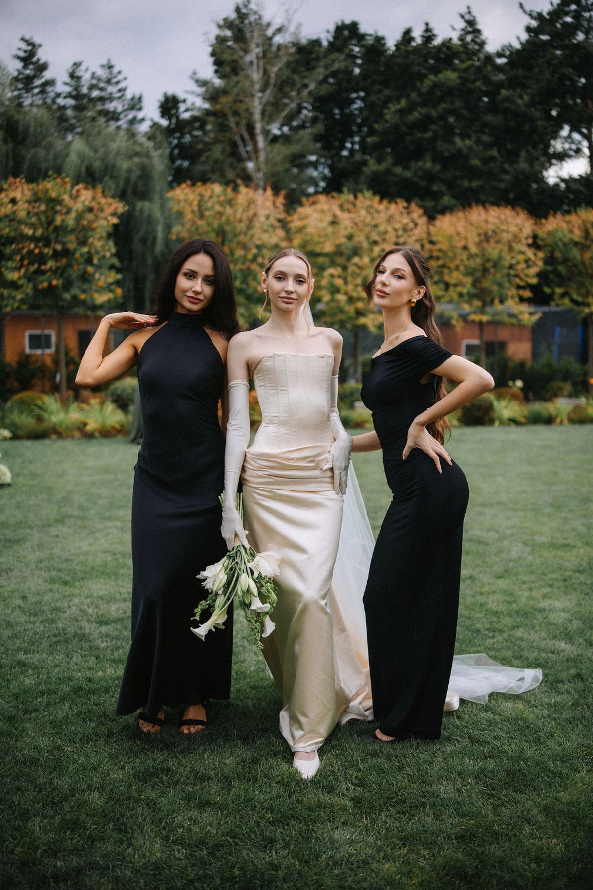 Three women pose together on a lawn wearing elegant dresses, with two in black gowns and one in a cream wedding dress.