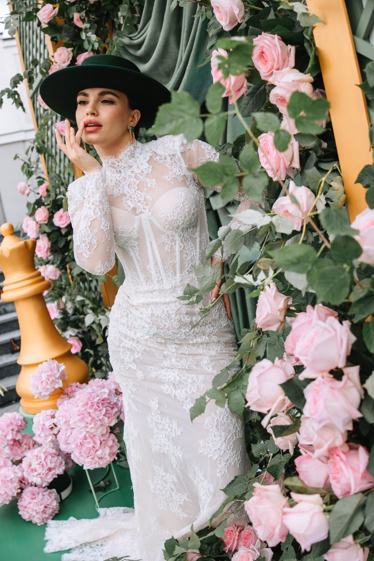 A romantic bridal portrait surrounded by pink roses and greenery against a yellow pillar in an elegant white lace gown.
