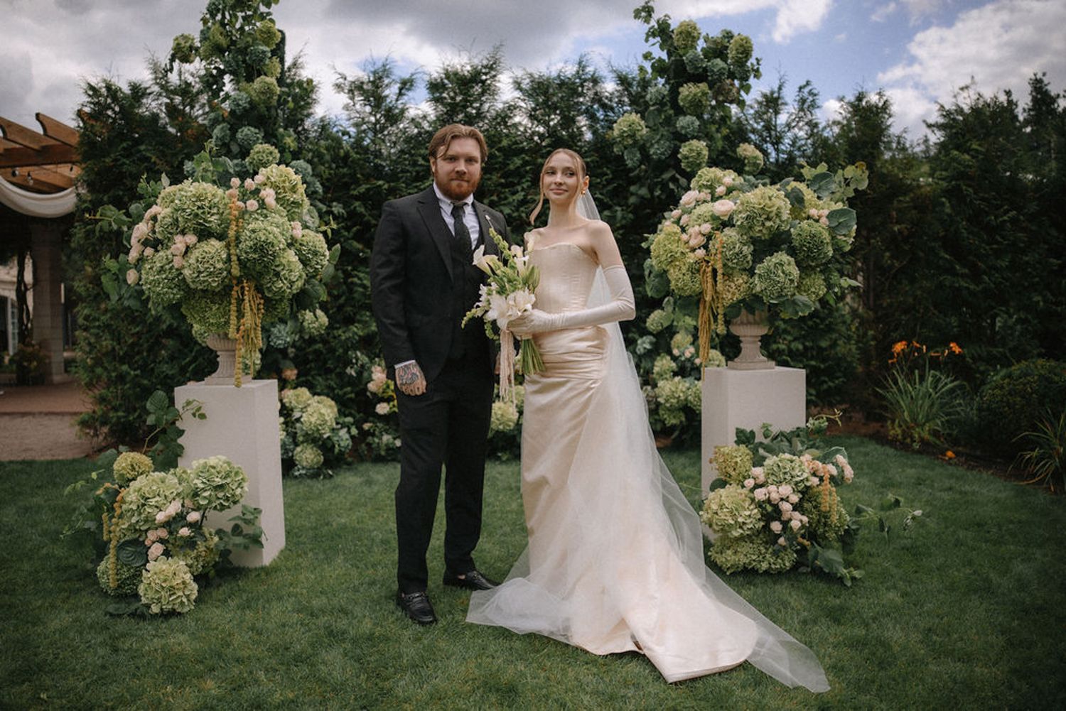 Elegant outdoor wedding ceremony setup with floral arrangements and guests gathering under cloudy summer skies.