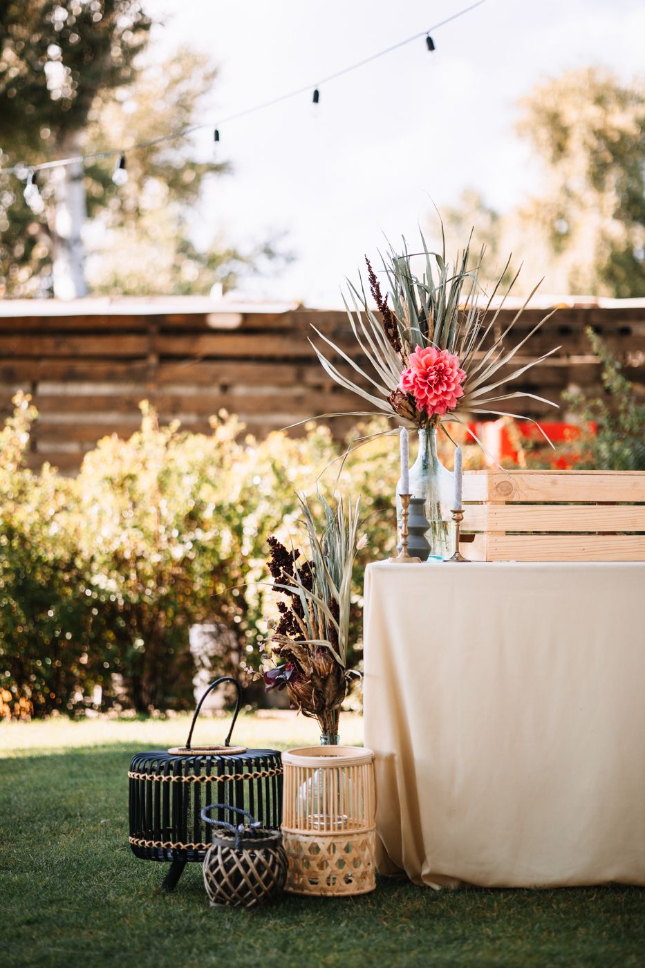 Outdoor garden party decor with woven baskets, string lights, and pink floral centerpiece on table with white tablecloth.