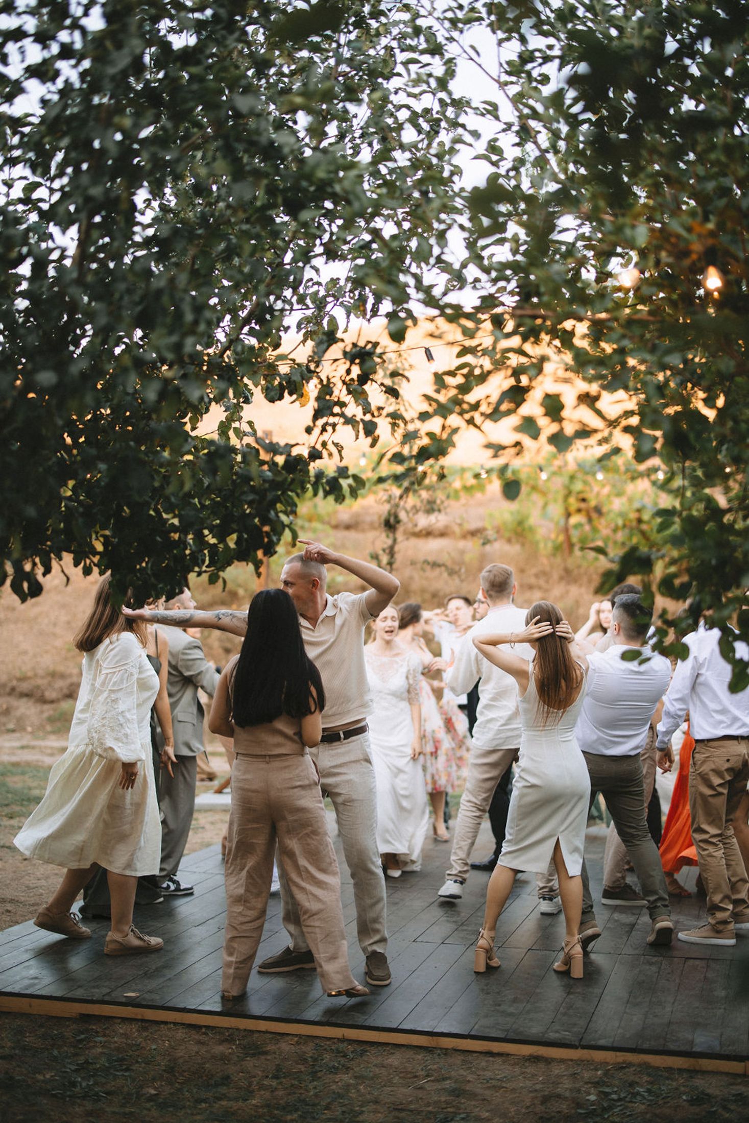 Wedding guests dance together in coordinated white and beige outfits on an outdoor dance floor at sunset.