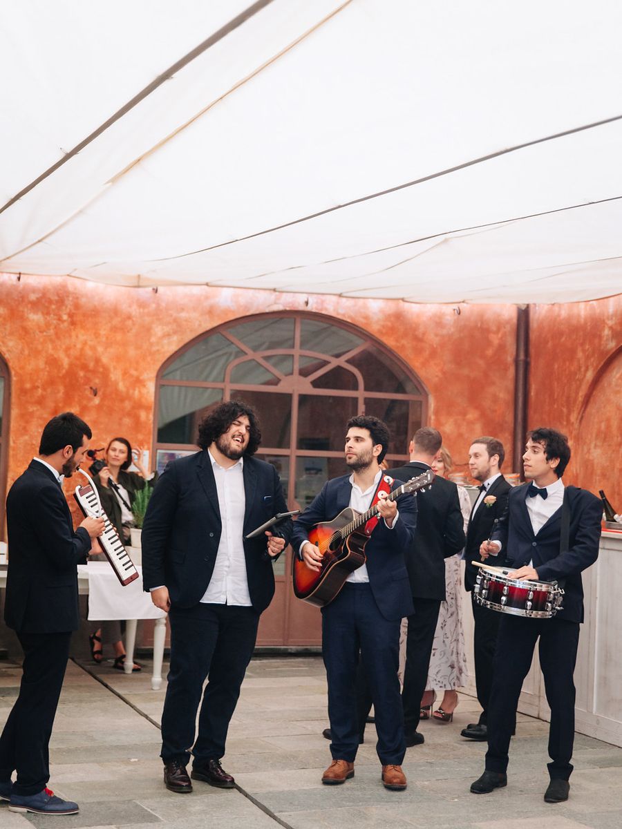 A musical band performs with guitars and drums against a rustic terracotta wall with an arched window.