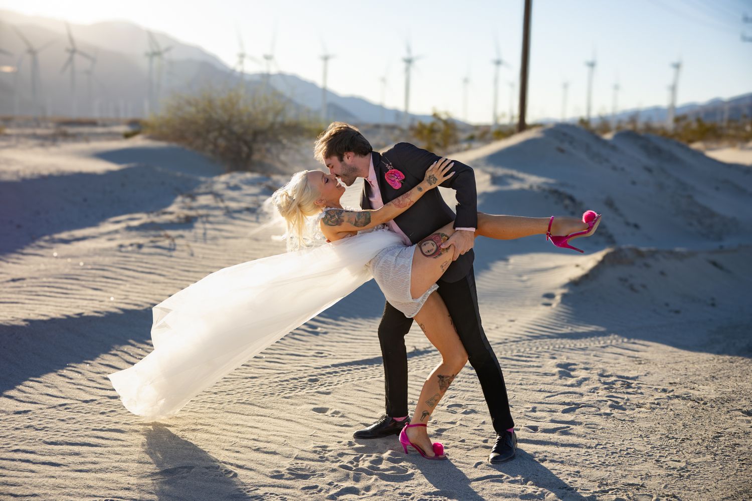 Couple sharing a romantic dip dance pose in a desert landscape with wind turbines, bride in white gown and pink heels.
