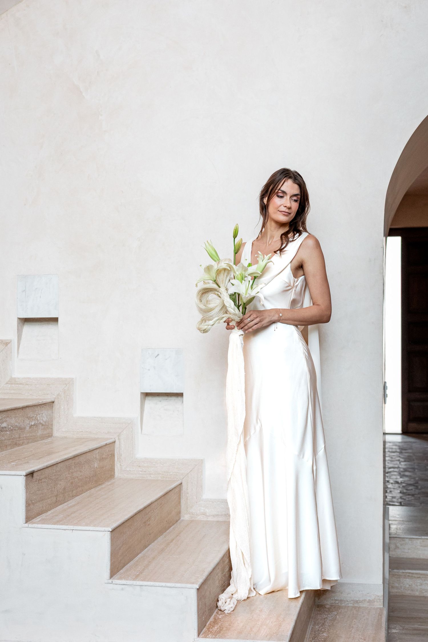 Bride in white satin gown holding white lily bouquet on stairs in elegant minimalist interior.