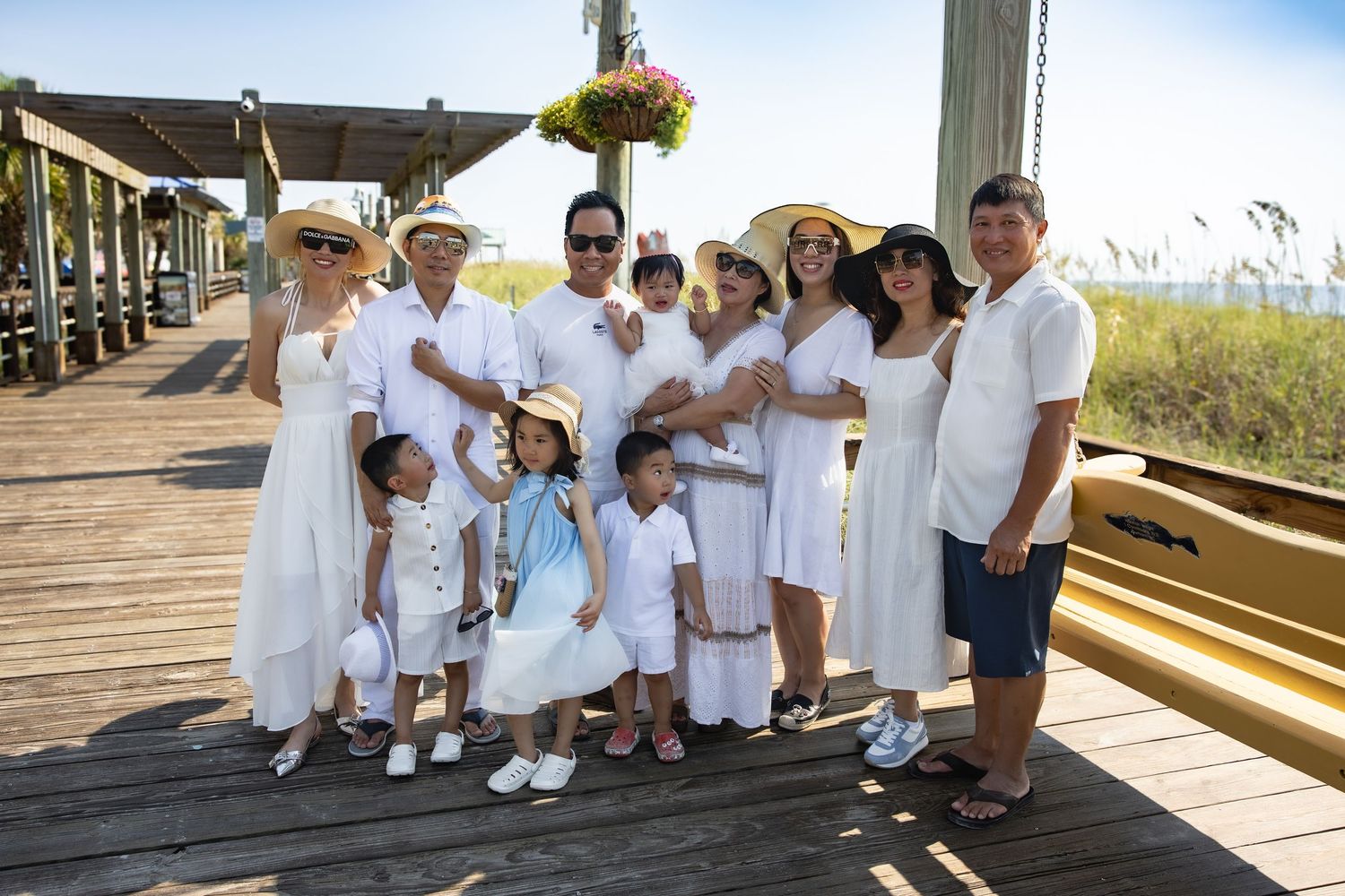 Group wearing coordinated white outfits poses together on a wooden boardwalk by the beach.