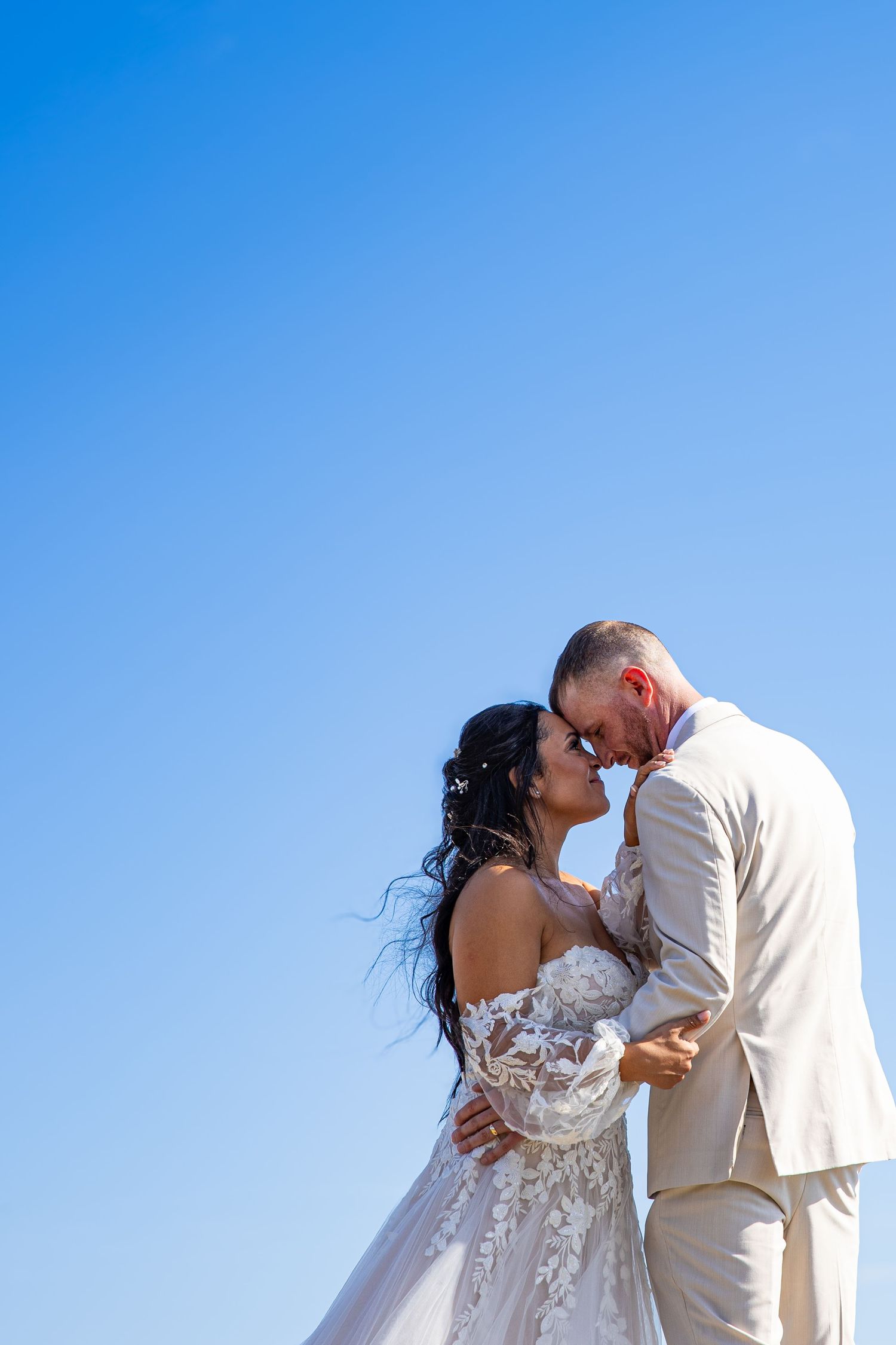 A romantic wedding couple shares a kiss against a bright blue sky on their special day.