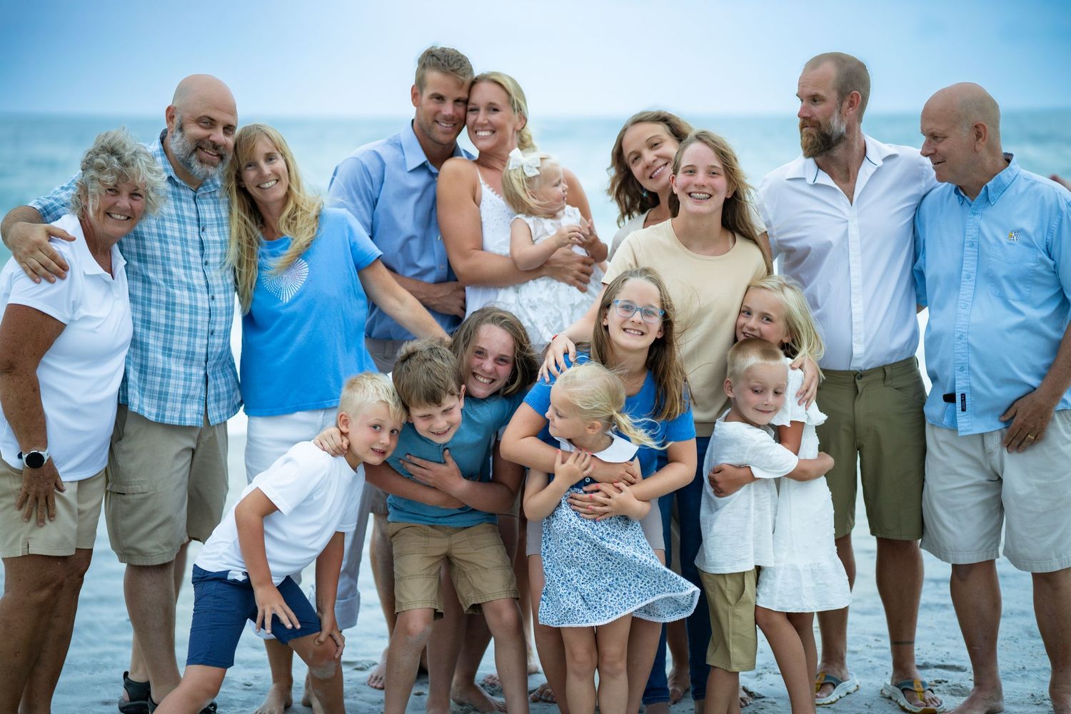 Large group poses together on beach wearing casual summer attire with ocean in background.