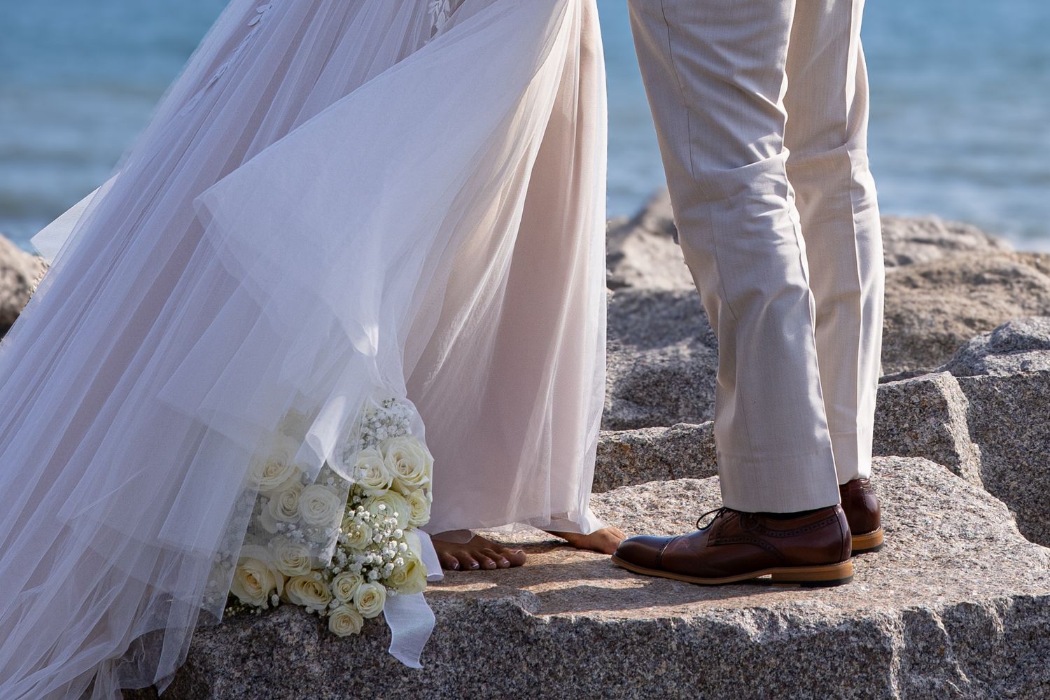 Wedding dress and groom's shoes on coastal rocks with white floral bouquet.