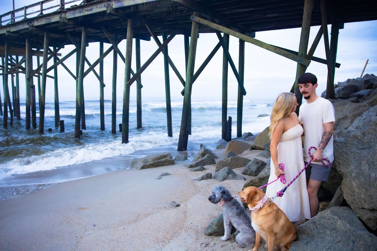 Couple stands near pier with their dog on rocky beach.