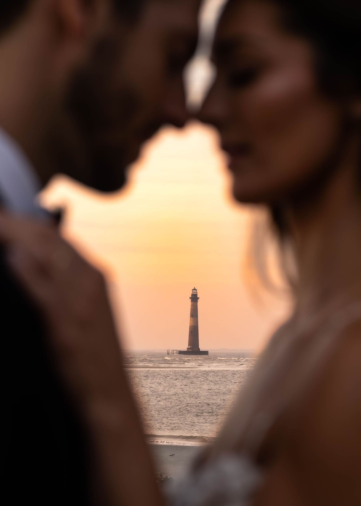 Lighthouse silhouetted against sunset sky framed between two figures in intimate moment.