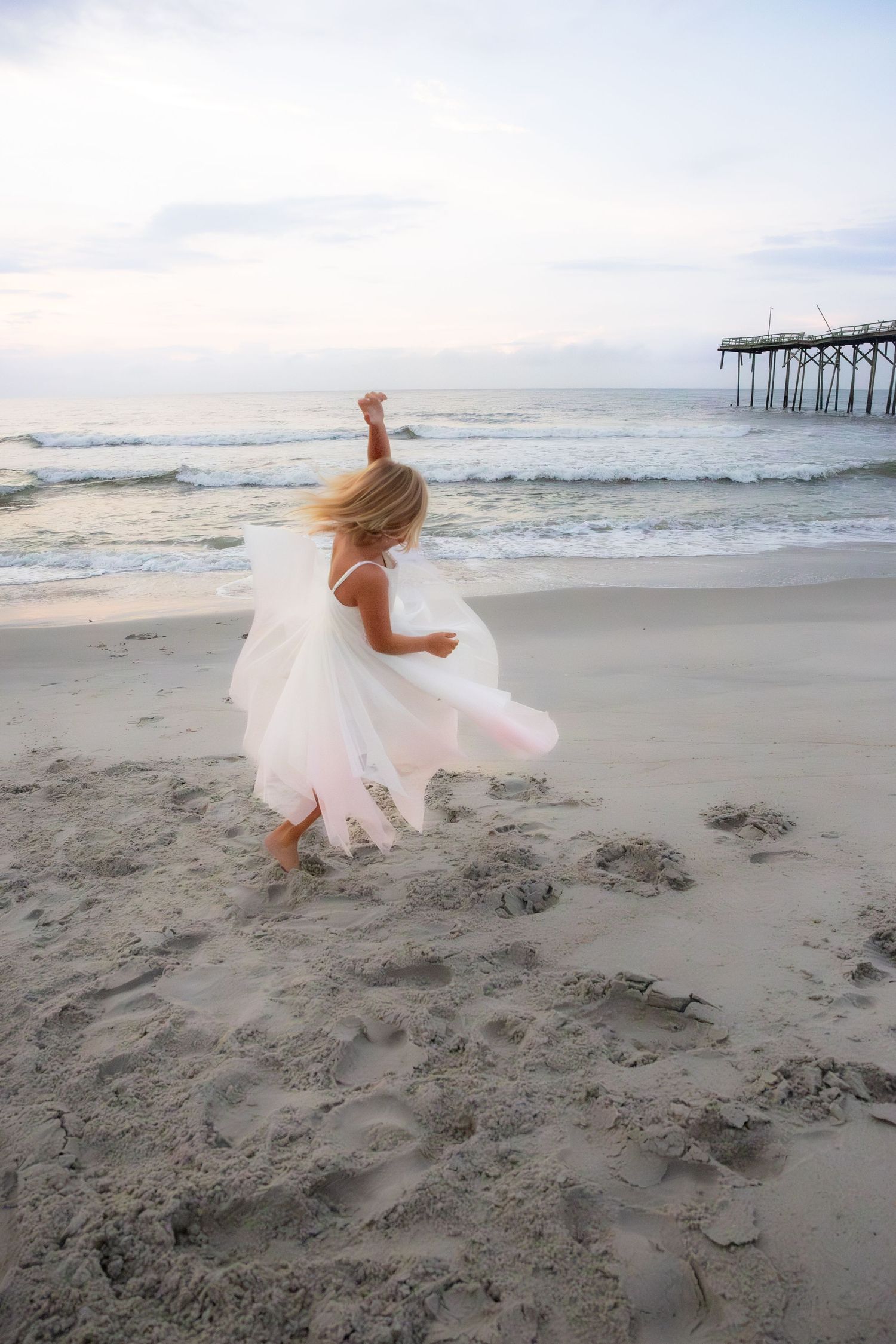 Flowing white dress twirling on a beach at sunset near a wooden pier.