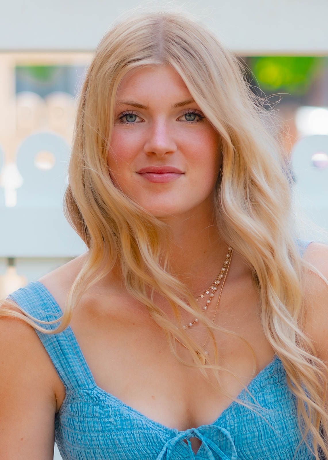 Young woman with long blonde wavy hair wearing a blue smocked top and delicate gold necklace outdoors.