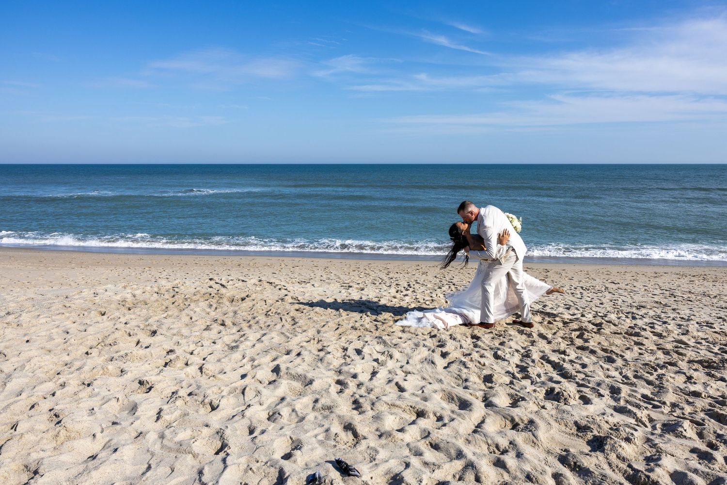 Bride in white wedding dress bending down on a sandy beach with ocean waves and blue sky in the background.