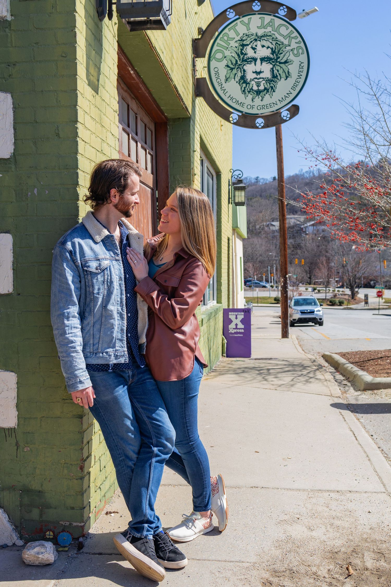 A couple poses affectionately outside a coffee shop with green brick walls.