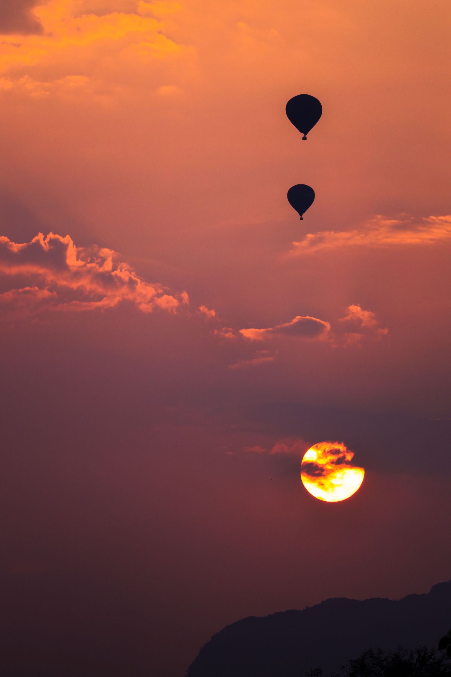 Two hot air balloons silhouetted against a vibrant orange and purple sunset sky with a glowing sun and dramatic clouds.