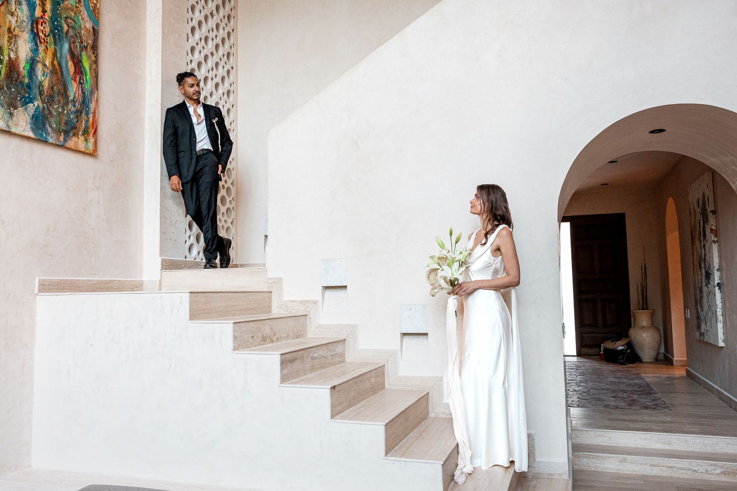 Bride in white gown holding bouquet looks up at groom in suit on elegant staircase during wedding ceremony.