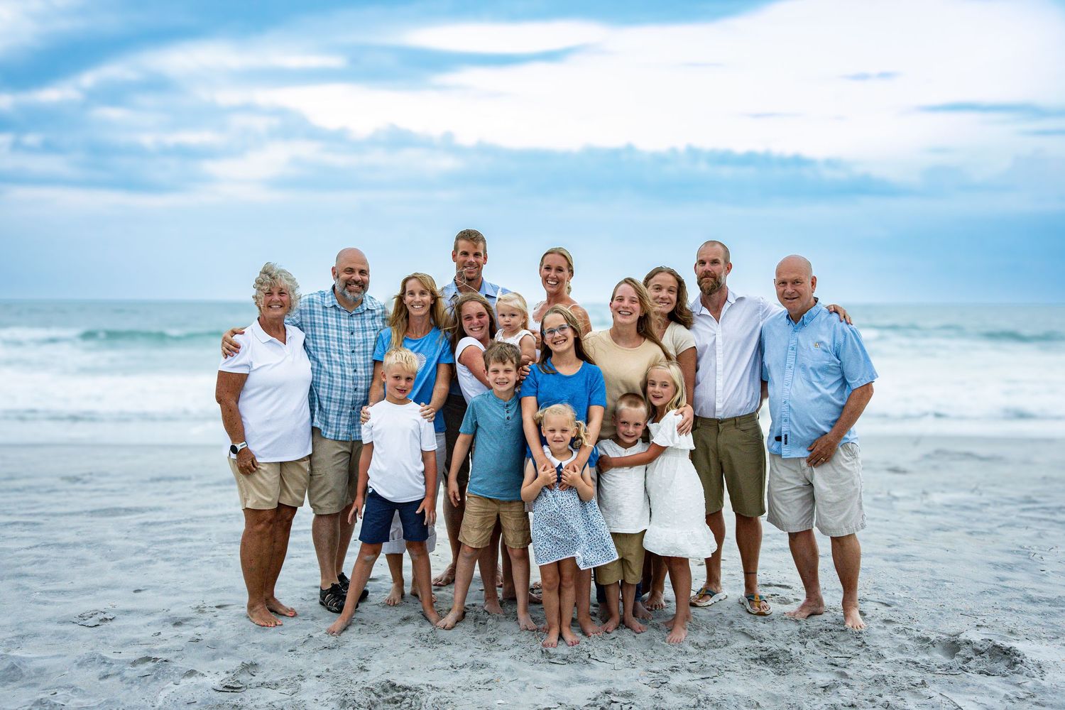 Extended family group poses together on beach with ocean waves and blue sky in background.