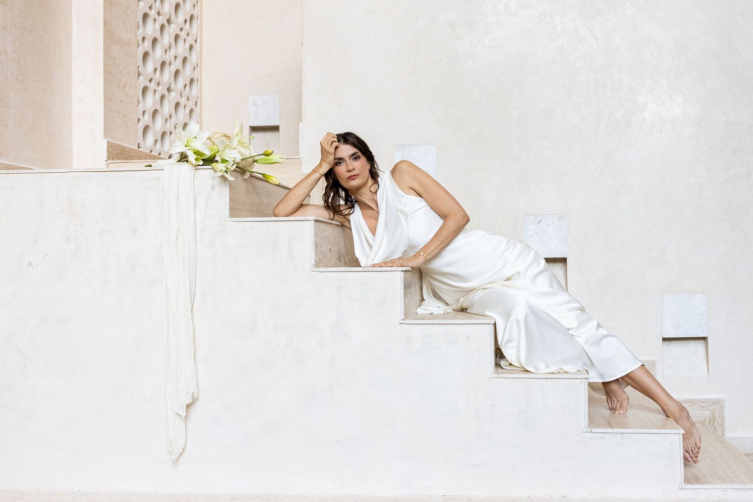 Woman in white dress posing on white stairs holding yellow flowers in a bright Mediterranean setting.