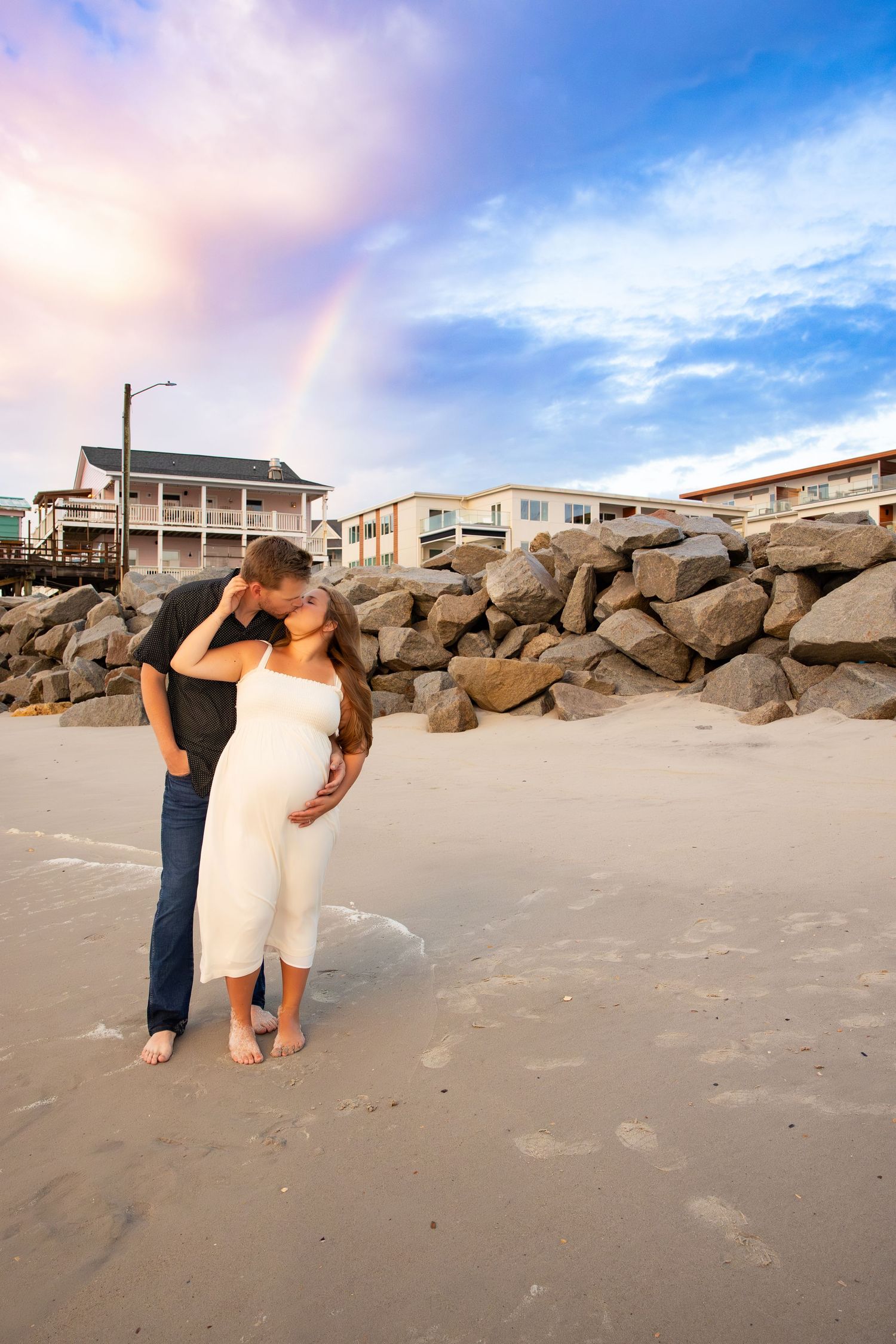 Couple embracing on beach with rainbow and coastal homes in background.