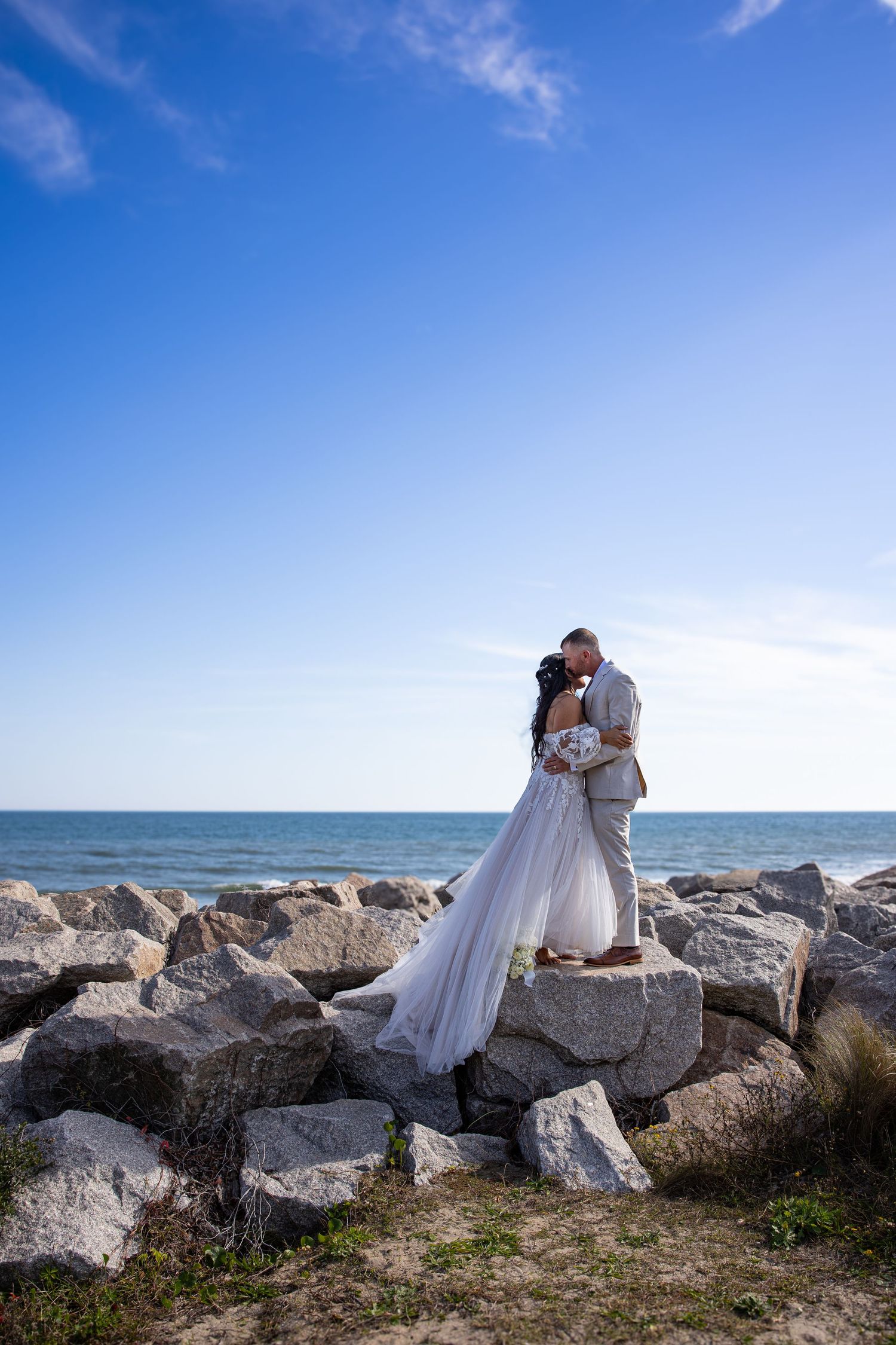 Romantic wedding couple embracing on rocky beach shore with bright blue sky.