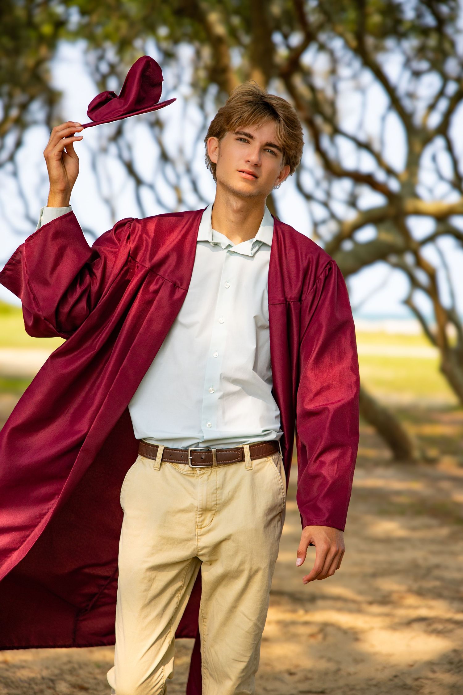 Young man in burgundy graduation gown and cap posing outdoors under trees wearing khaki pants and white shirt.