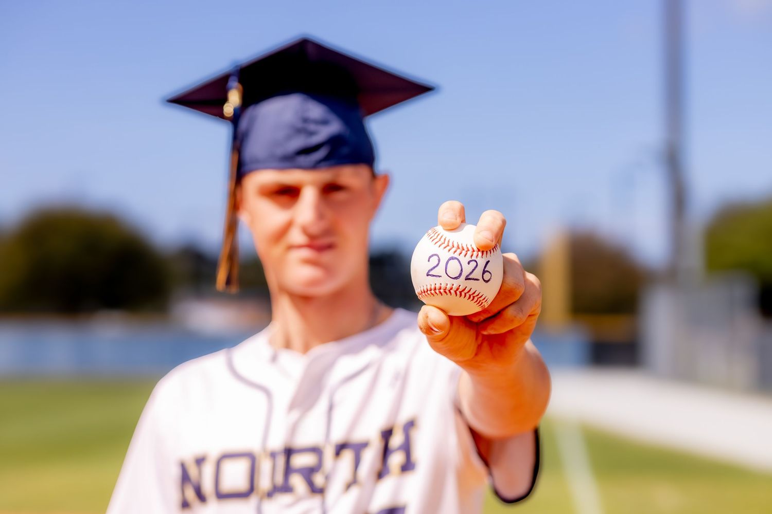 High school senior in graduation cap holds baseball marked 2026, wearing North jersey on baseball field.