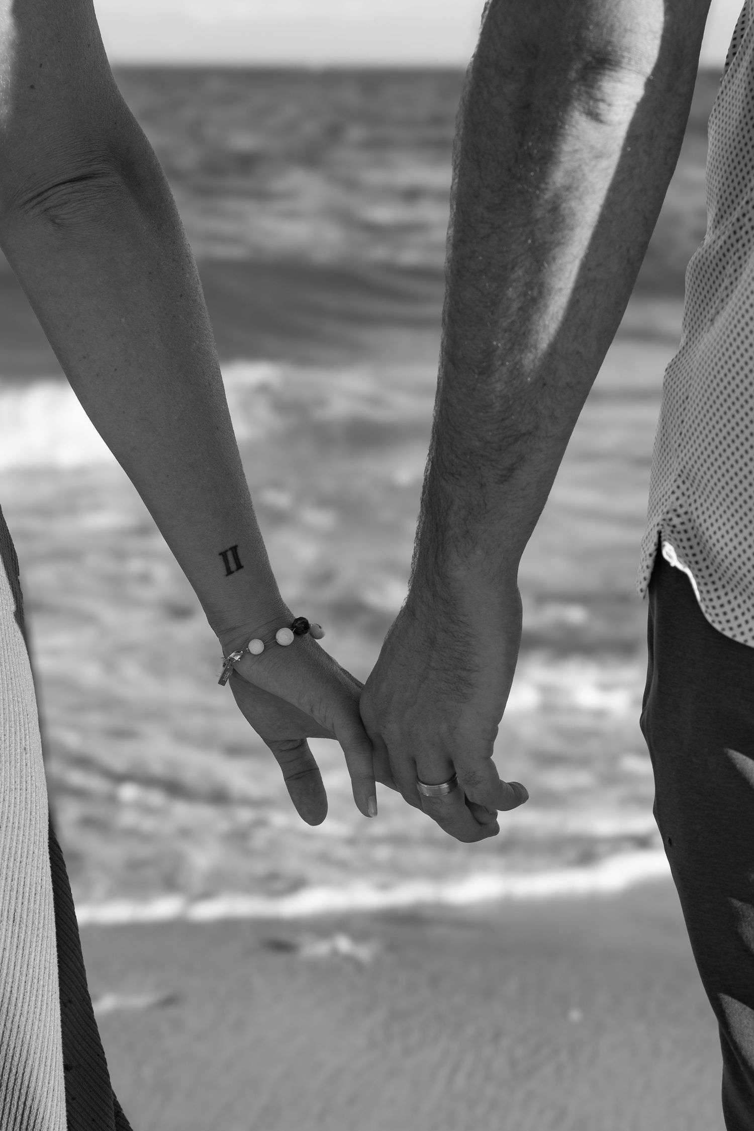 Black and white close-up of hands holding each other against an ocean backdrop.