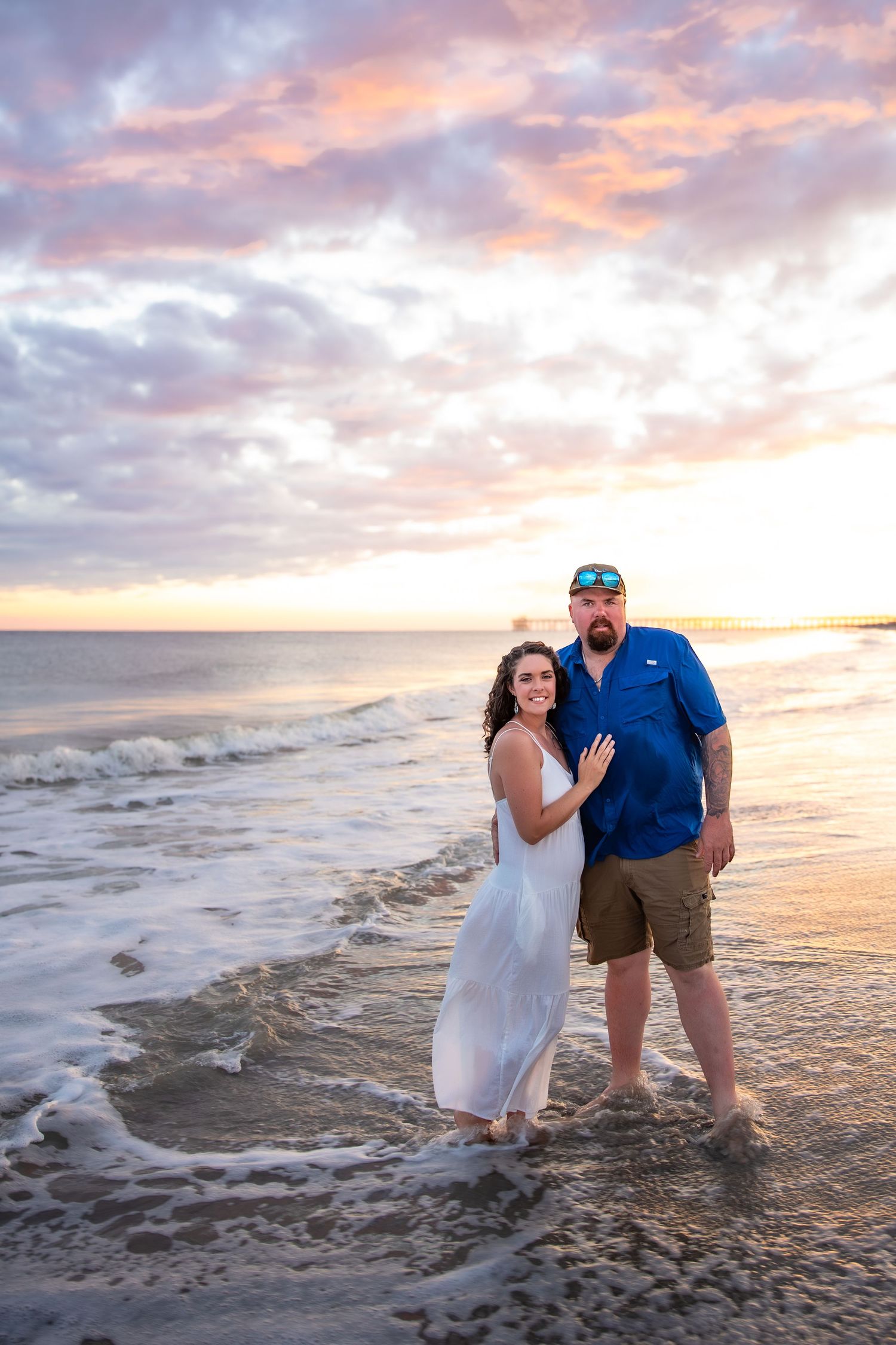 Couple stands together in ocean waves during golden sunset at the beach.