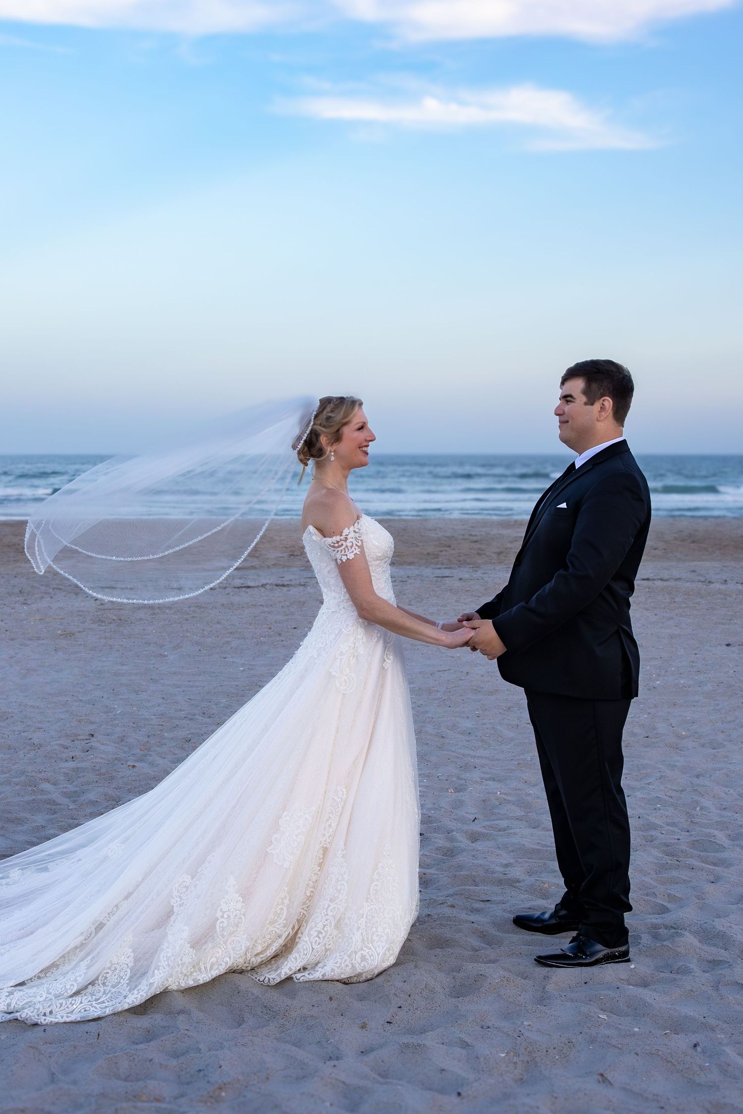 A bride's flowing white dress trails beautifully across the beach sand during sunset portraits.