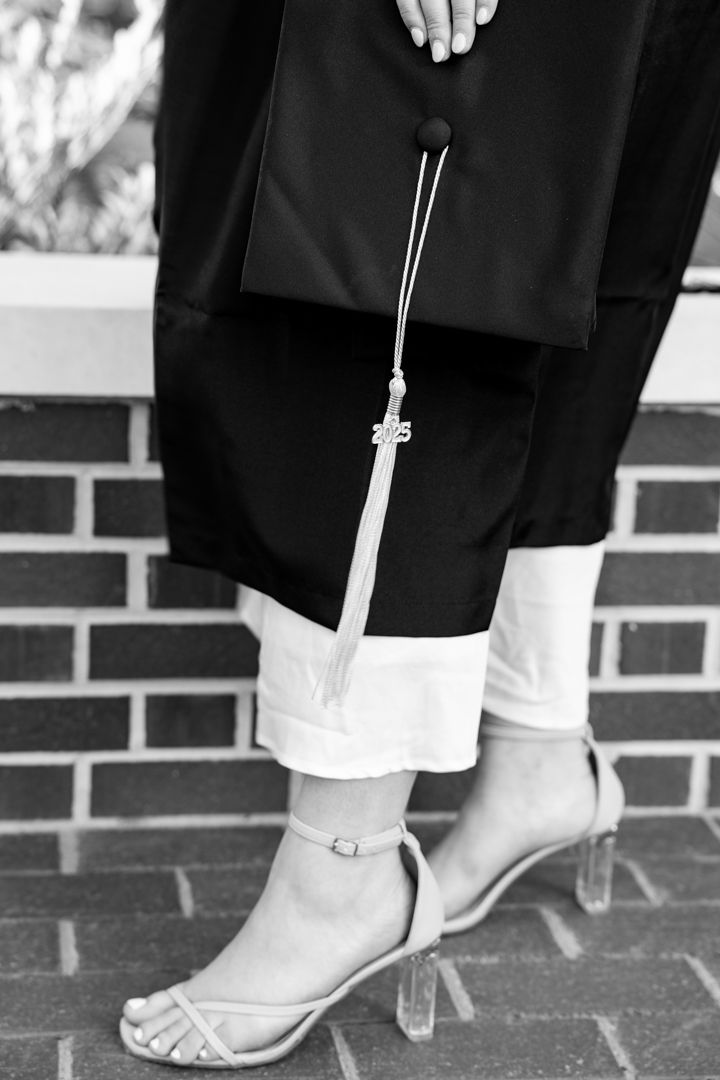 Black and white photo of a graduate holding cap with tassel, wearing white pants and strappy heels on brick steps.