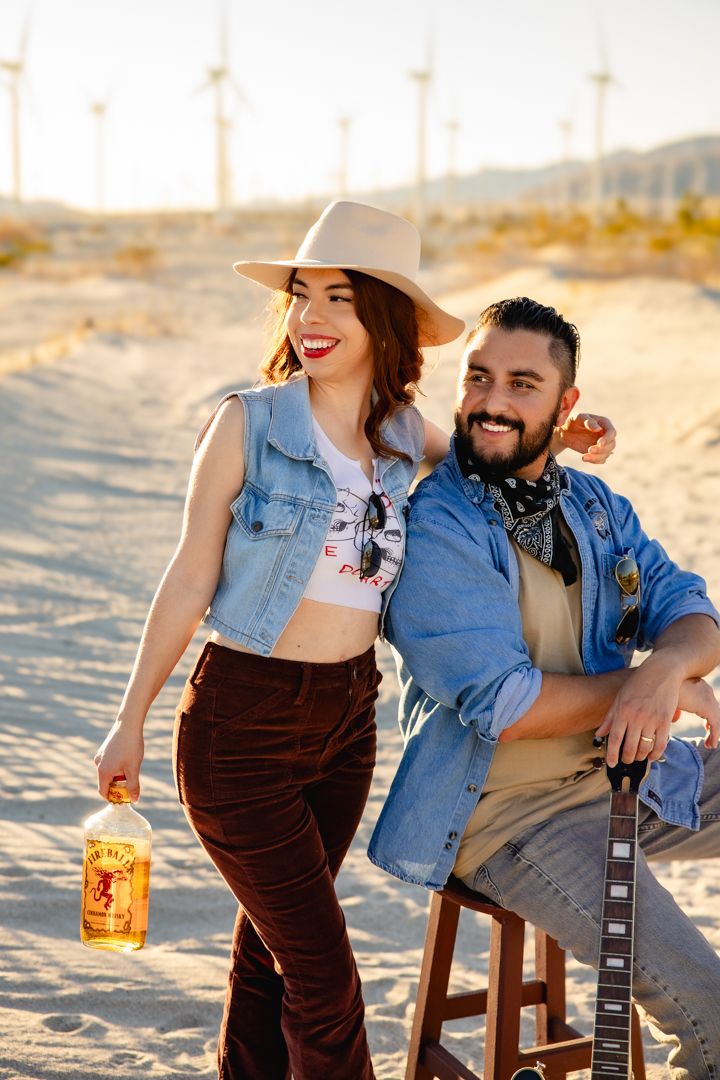 Couple enjoying a beach photoshoot with guitar and drinks at sunset.