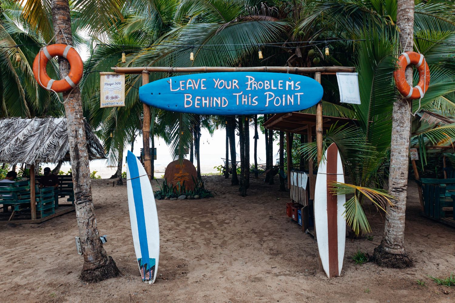 Tropical beach entrance with surfboards and sign reading Leave Your Problems Behind This Point framed by palm trees.