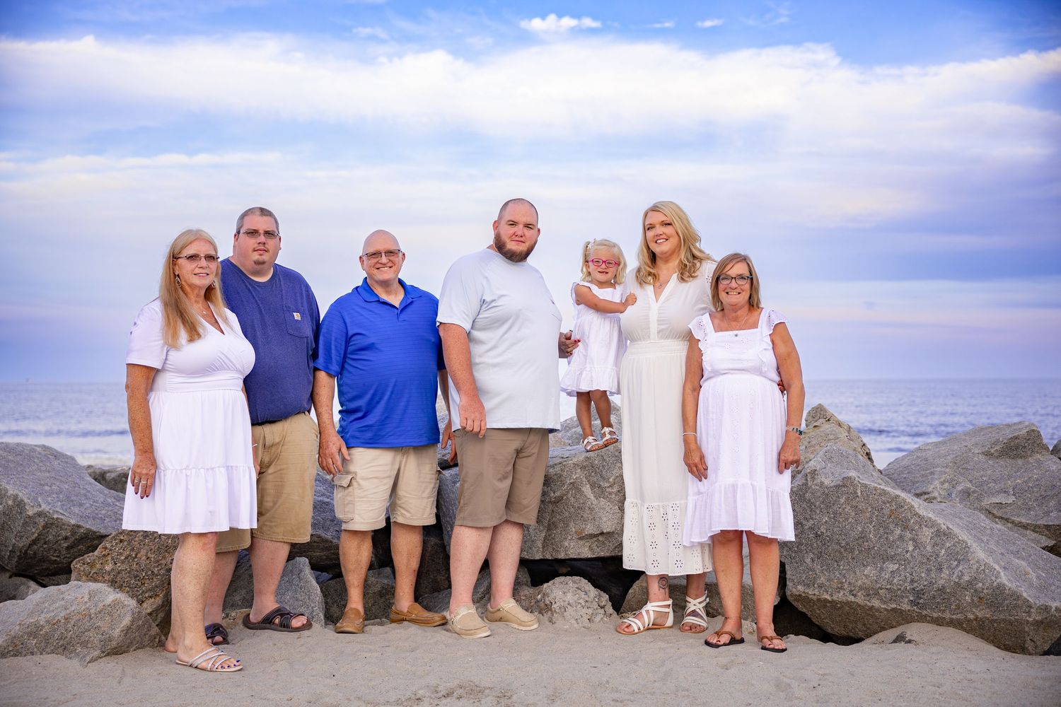 A group poses together on rocky coastal jetty with ocean and cloudy sky in background.