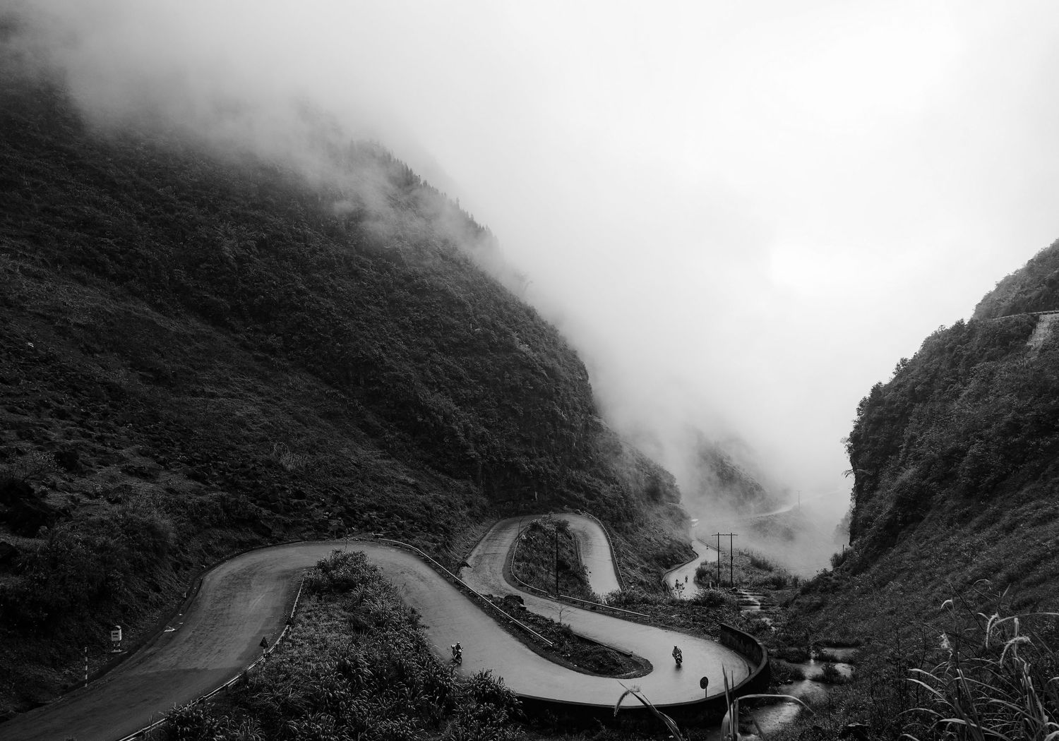 Black and white aerial view of a winding mountain road with hairpin turns shrouded in misty fog.