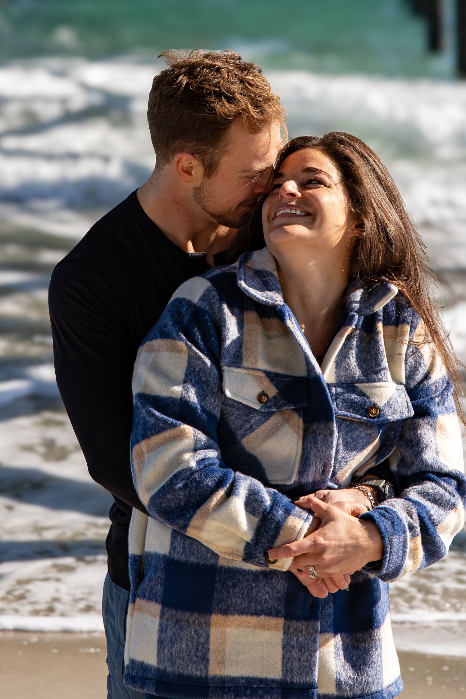 A couple embraces on a beach wearing a blue and white plaid blanket as waves crash behind them.