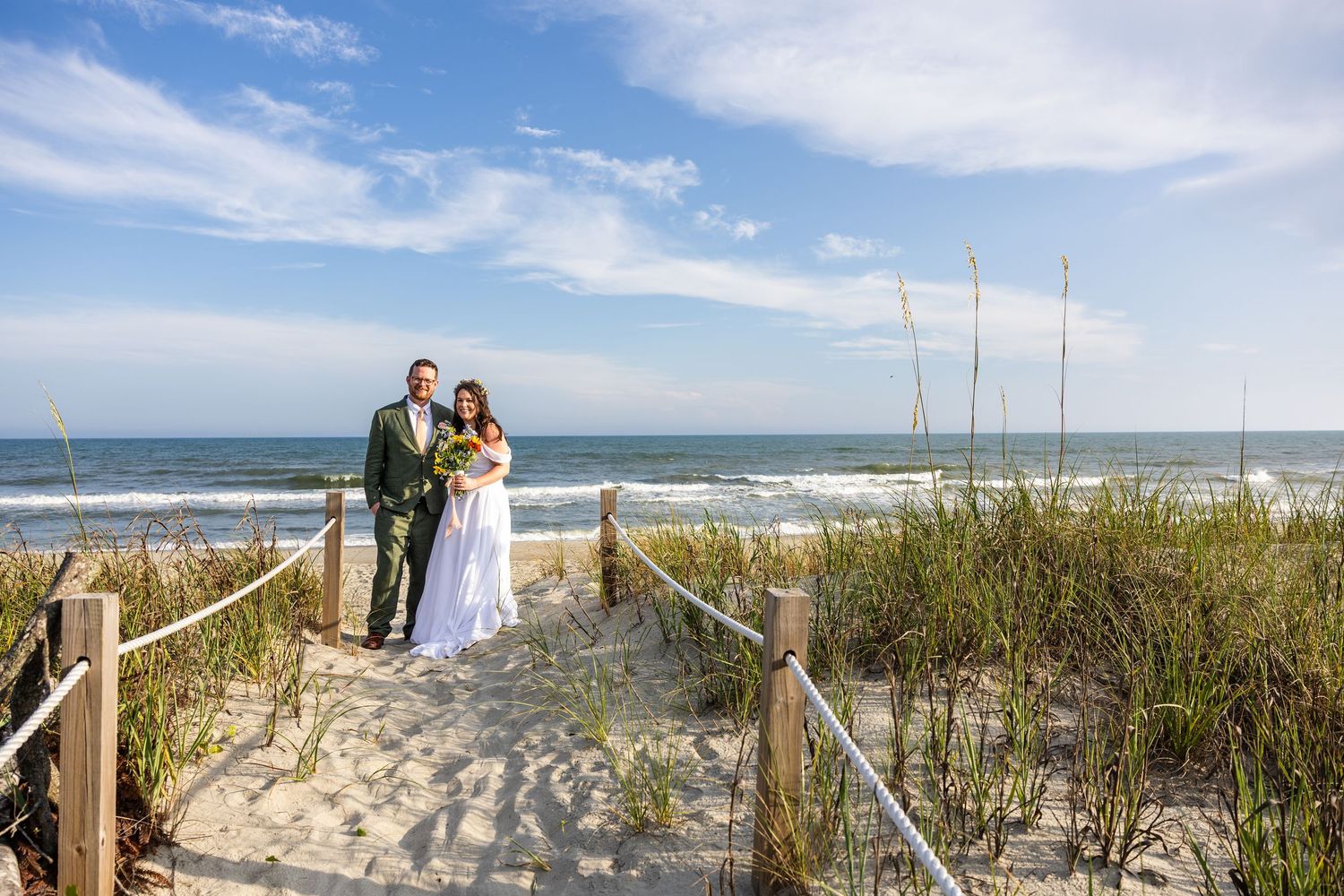 Bride and groom pose on sandy beach path with sea oats, ocean waves, and blue sky backdrop on their wedding day.