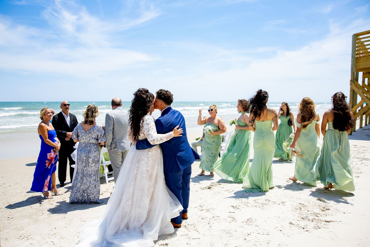 Bride and groom embrace on a sunny beach wedding with bridesmaids in mint green gowns and guests celebrating nearby.