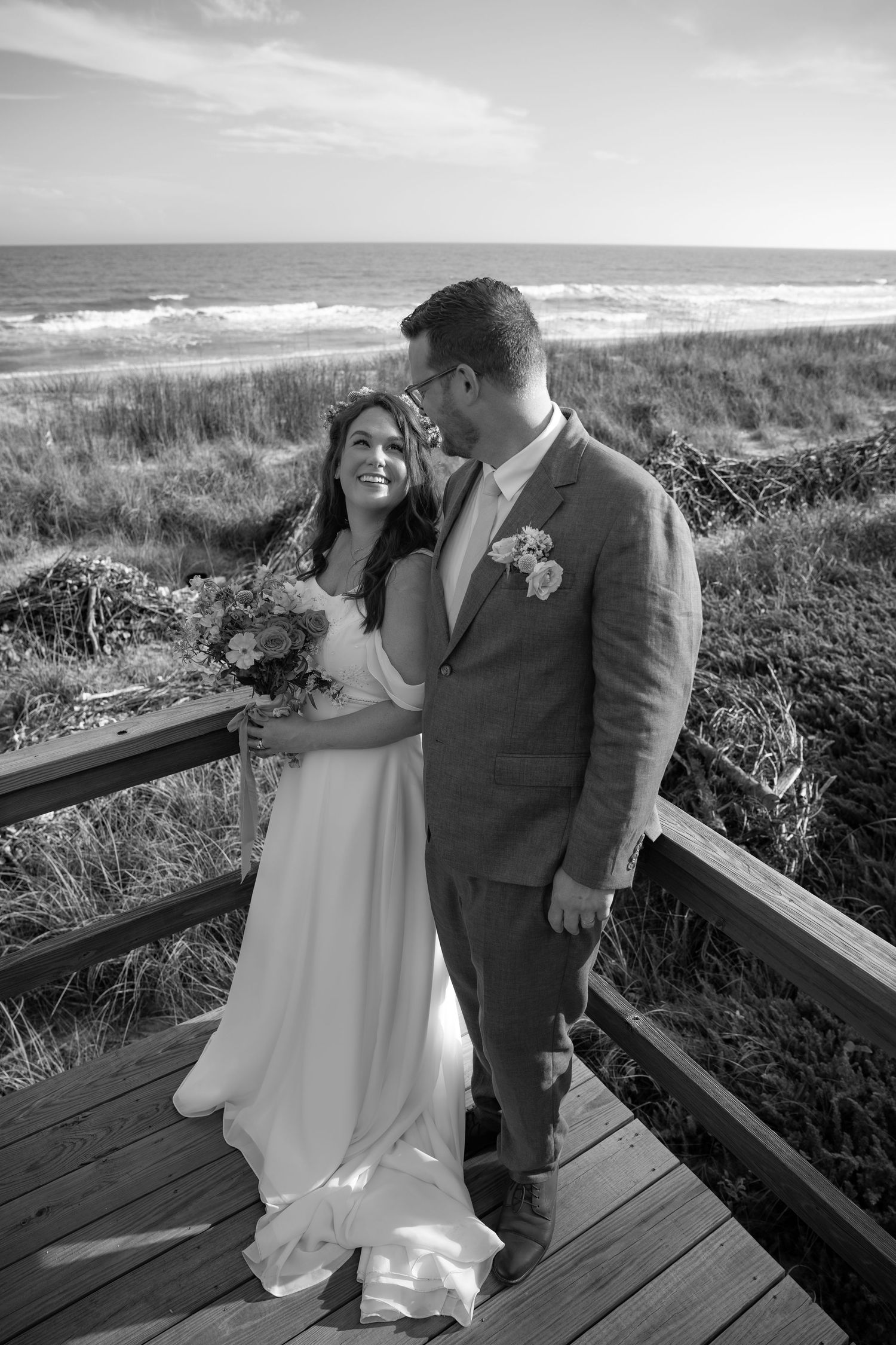 Black and white photo of a smiling bride and groom embracing on a coastal boardwalk with ocean waves in the background.