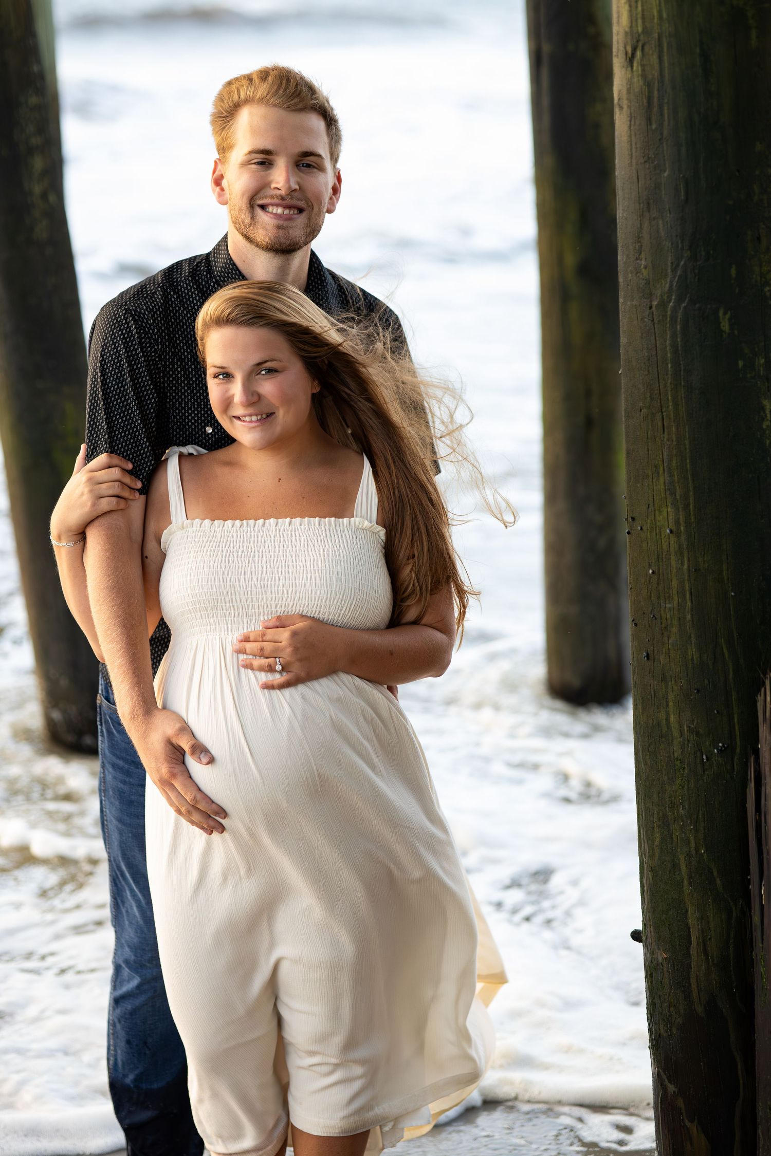 A romantic couple embraces by wooden pier posts at the beach with waves in the background.
