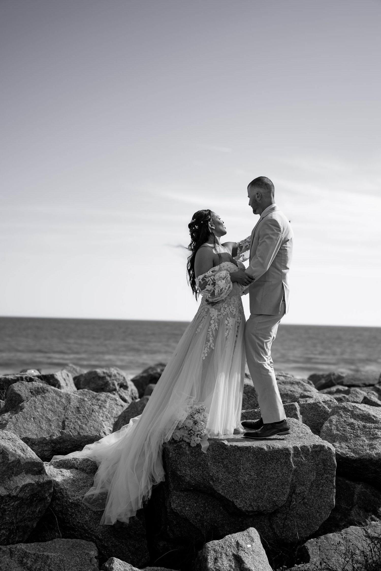 A romantic black and white photo of a couple standing together on coastal rocks by the ocean.