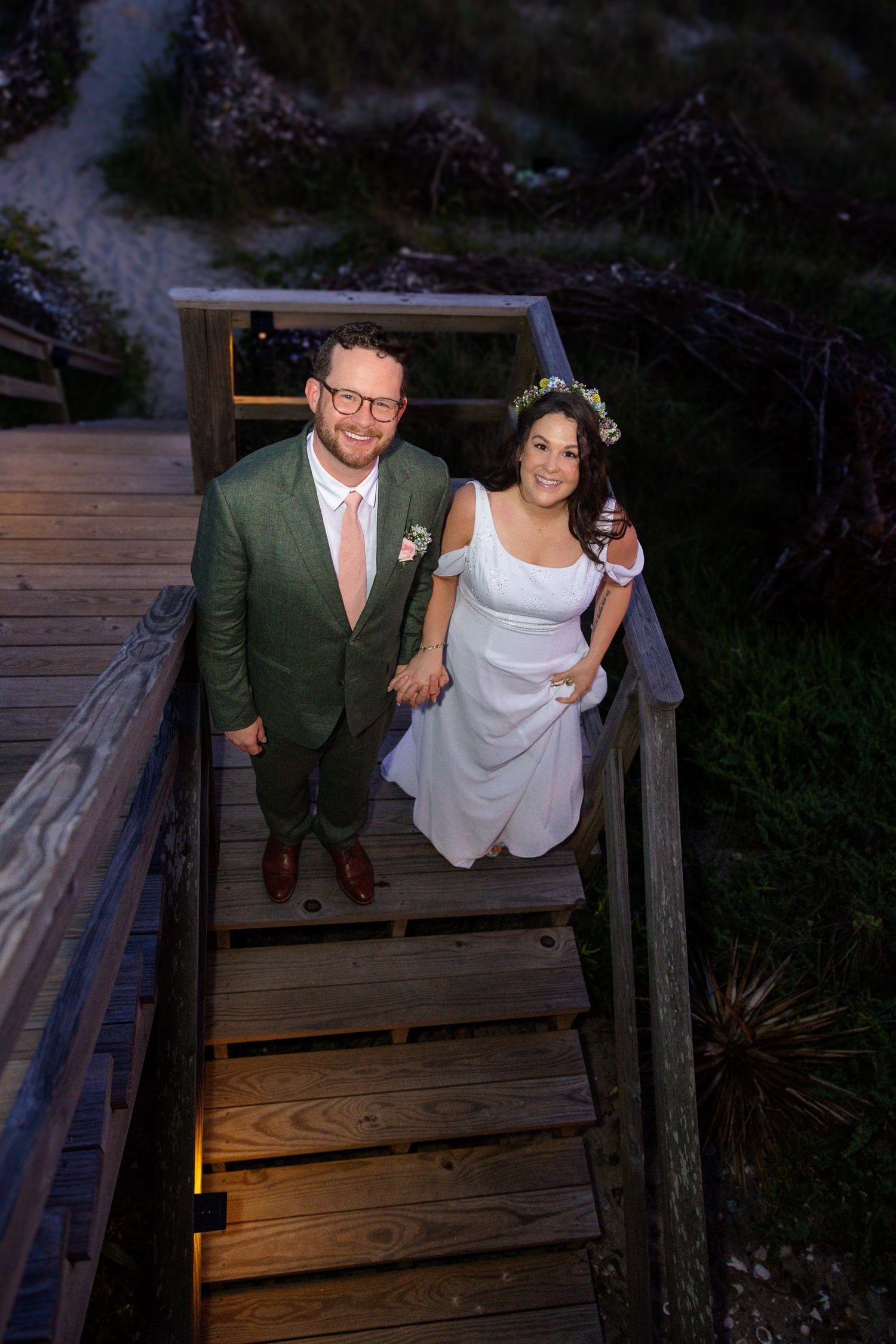 Bride in white dress and groom in green suit hold hands on wooden boardwalk steps at dusk wedding photo.