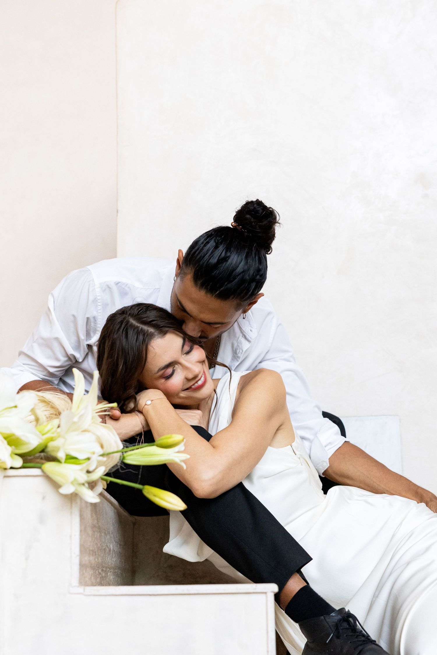 Couple sharing a tender embrace with white lilies on a table in a bright minimalist room.