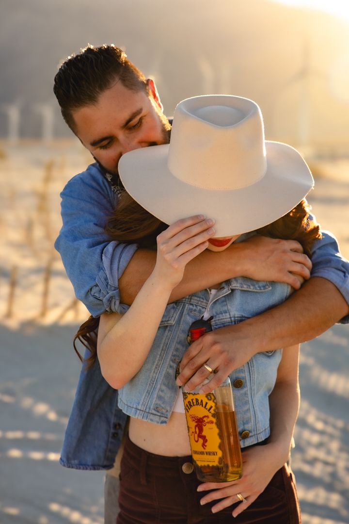 A romantic couple embraces on a beach at sunset while holding a bottle of Fireball whiskey.