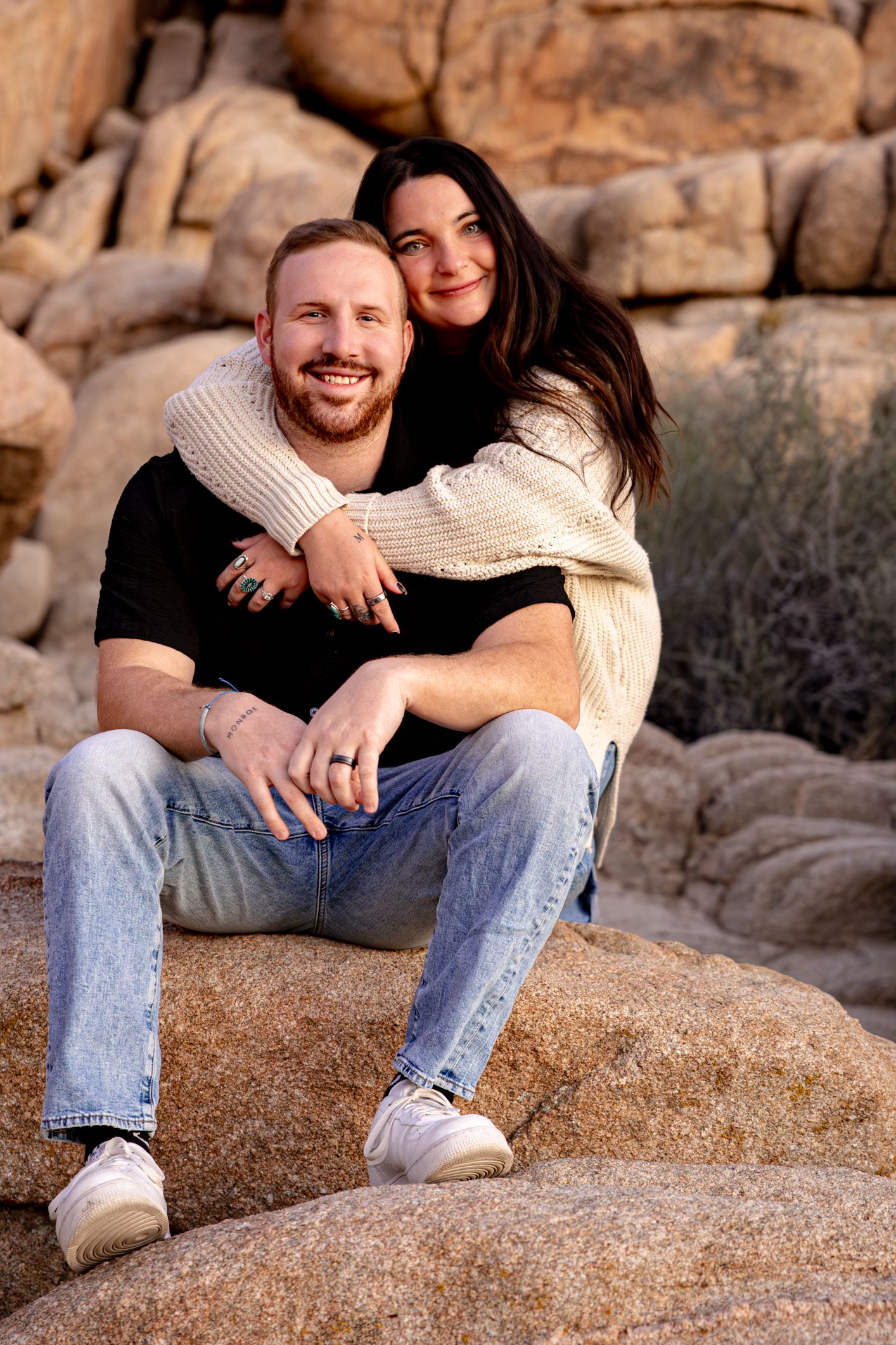 A couple sits together on coastal rocks during a romantic outdoor photoshoot at golden hour.