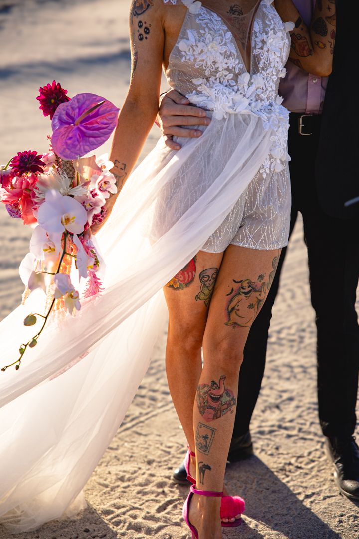 Close-up of a wedding dress with floral lace details and visible tattoos on the wearer's legs.