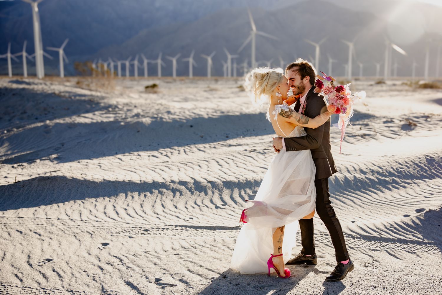 Bride and groom embracing in sandy desert with wind turbines backdrop in Palm Springs wedding photo.