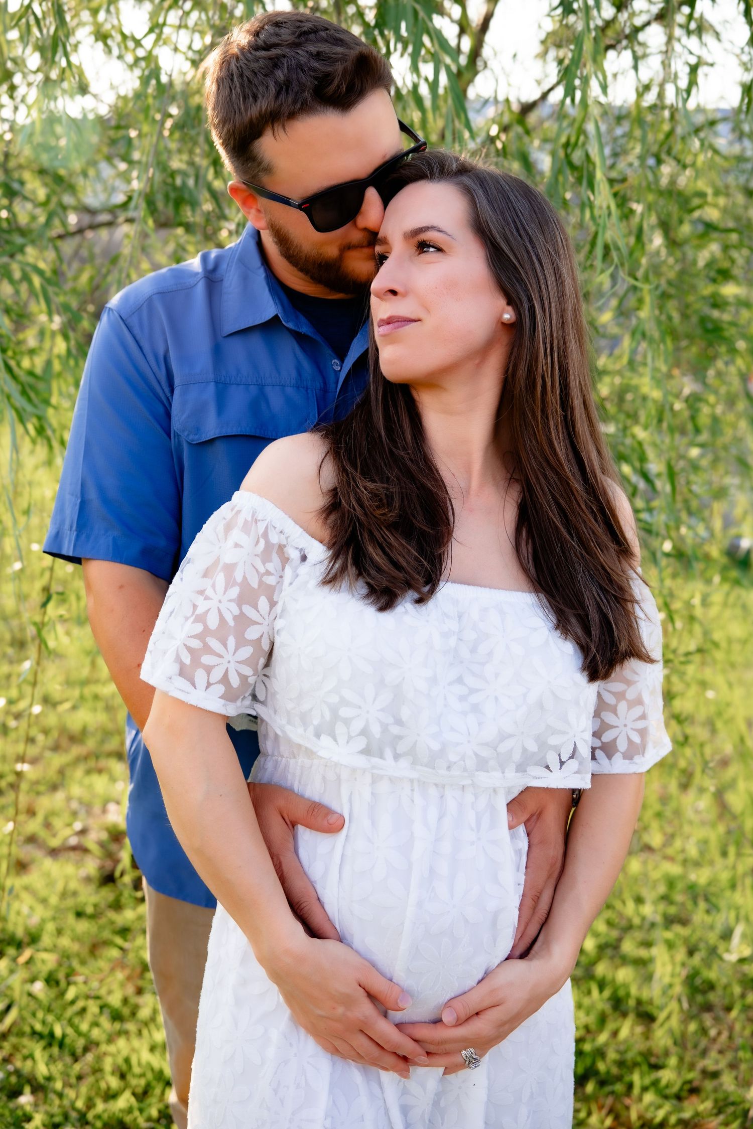A couple in a blue shirt and white lace dress embrace lovingly outdoors among green foliage.