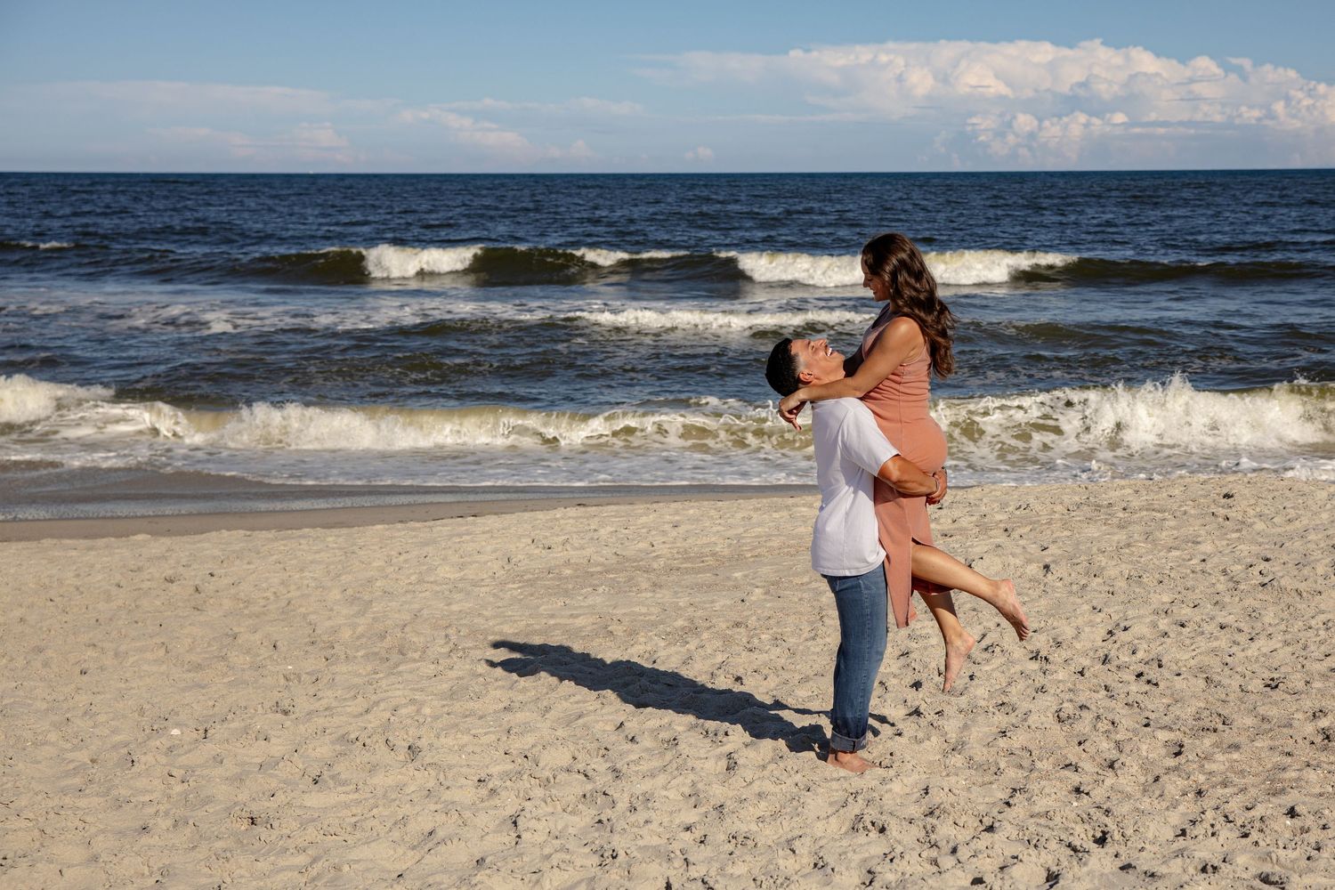 Two people embrace on a sandy beach with ocean waves and white clouds in the background.