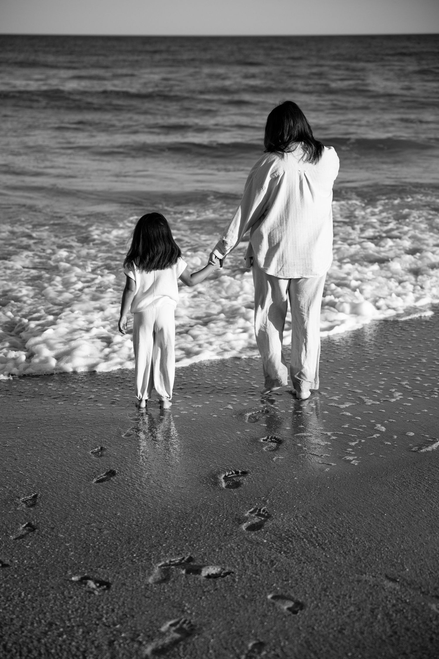 Black and white image of two people walking along beach leaving footprints in wet sand.