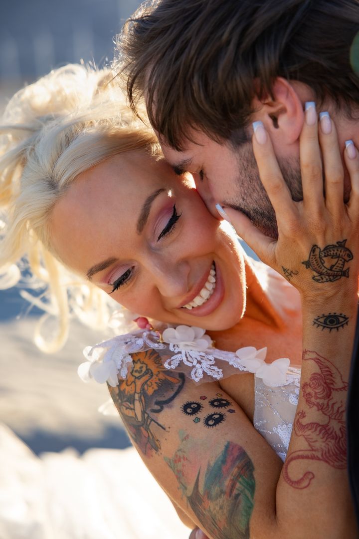 A smiling tattooed blonde bride being kissed on the cheek by her groom on their wedding day.